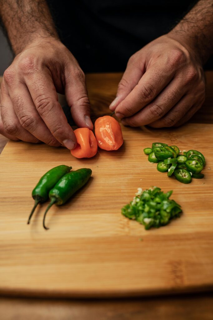 Hands preparing fresh jalapenos and habanero peppers on a wooden chopping board.